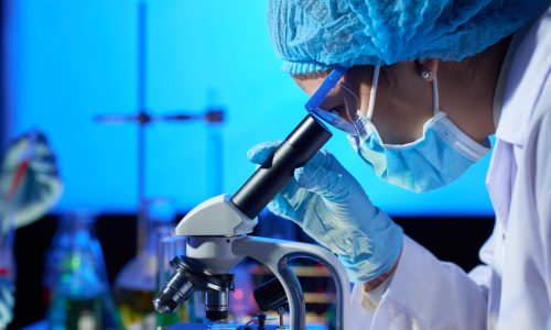 Profile view of concentrated Asian microbiologist examining sample with help of modern microscope while wrapped up in work at dim laboratory