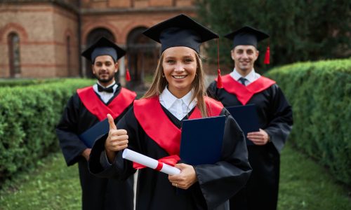 Graduate female shows like with her friends in graduation gowns holding diploma and smiling at camera.