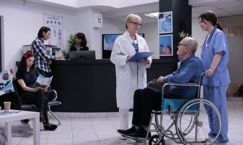 Retired person using wheelchair talking with senior doctor for appointment while being helped by professional nurse in private clinic. Older man living with disability in busy hospital reception.